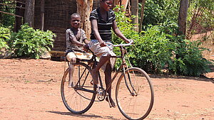 Zwei Kinder in Malawi fahren gemeinsam lachend auf einem Fahrrad über einen staubigen Platz