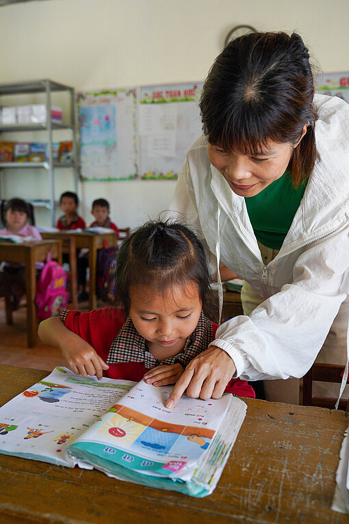 Lehrerin unterstützt eine junge Schülerin beim Lesen in einer Grundschule in Vietnam. Bildung für Mädchen als Motor für Veränderung.
