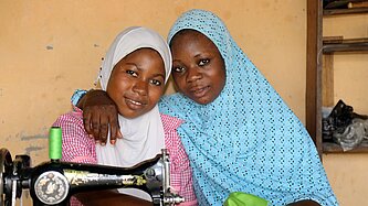 Zwei Mädchen mit Kopftuch sitzen Arm in Arm nebeneinander