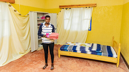 Eine Frau steht in einem Zimmer mit einem Bett und einem Schrank. Sie hält eine Packung Binden und ein Handtuch in ihren Händen. 