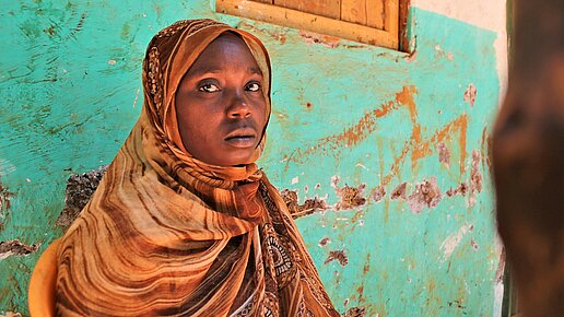 Eine verschleierte Frau sitzt auf einem Stuhl vor einer grünen Wand und schaut etwas verlegen in die Kamera