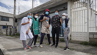 Fünf junge Erwachsene mit Masken posieren vor einer Bäckerei in Ecuador. Sie tragen Arbeitskleidung und zeigen Gesten der Zuversicht und Gemeinschaft.