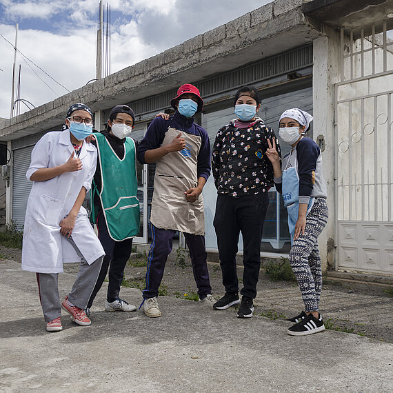 Fünf junge Erwachsene mit Masken posieren vor einer Bäckerei in Ecuador. Sie tragen Arbeitskleidung und zeigen Gesten der Zuversicht und Gemeinschaft.