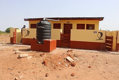 Ein kleines Haus hat zwei Eingänge, einer für Mädchen und einer für Jungen. Es sind Latrinen. Vor dem Gebäude steht ein Wassertank.