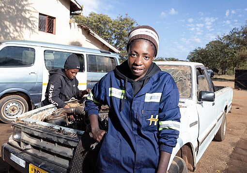 Eine junge Frau im Overall und mit Mütze sitzt auf ein Auto gelehnt