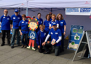 Mitglieder der Plan-Aktionsgruppe Köln posieren in blauen Plan-T-Shirts vor einem Infostand.