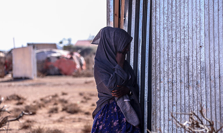 Eine junge Frau, die nicht zu erkennen ist, steht vor einer Wellblechhütte