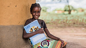 Ein Mädchen sitzt in Simbabwe an einer Wand aus Lehm, hält ein Plan International Heft und lächelt. Sie trägt ein buntes Kleid mit einem Streifenmuster und sitzt vor einer ländlichen Kulisse in Simbabwe.