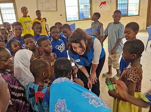Eine Frau mit einer blauen Weste mit Plan-Logo steht umringt von Kindern in einem Raum