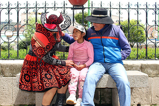 Jessica aus Chumbivilcas, Peru, sitzt mit ihrer Tochter und ihrem Mann auf einer steinernen Bank. Sie sehen sich an und lachen.