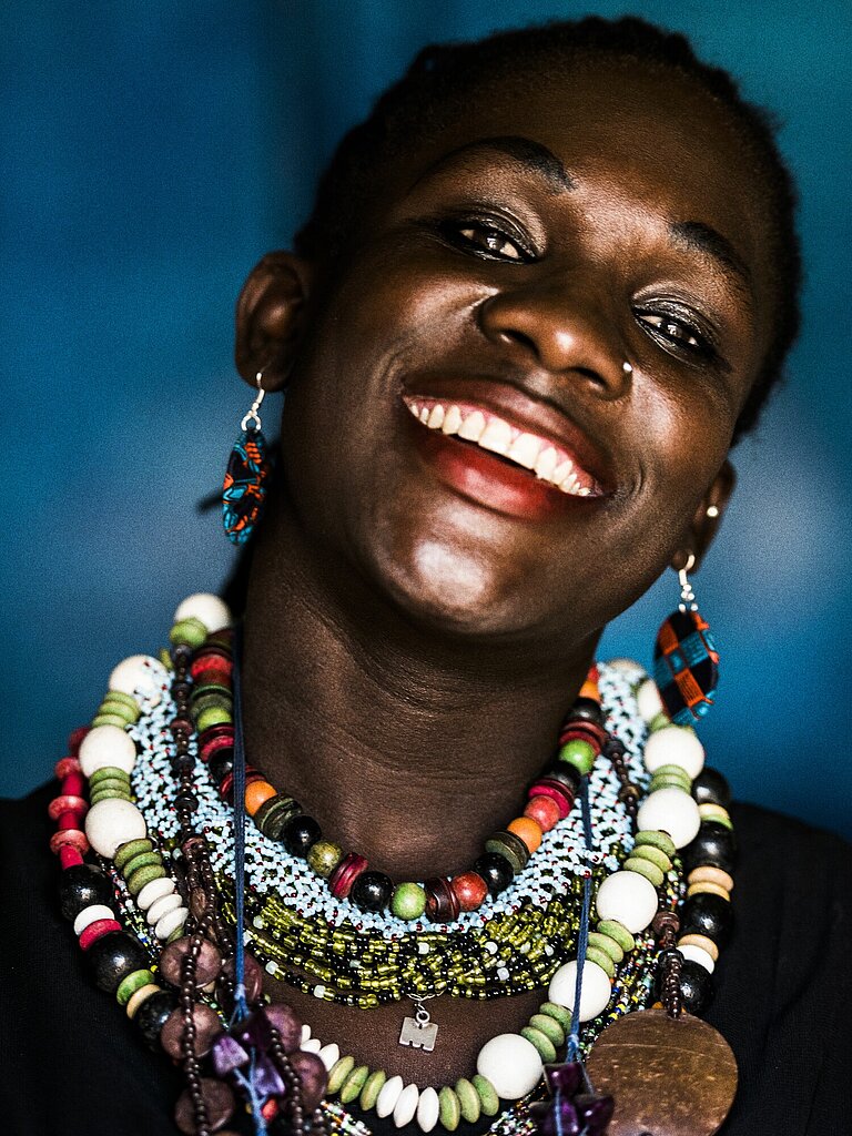 Portrait einer Schwarzen Frau, die den Kopf leicht seitlich gelehnt hat und lacht. Sie trägt viele verschiedene Ketten um den Hals. 