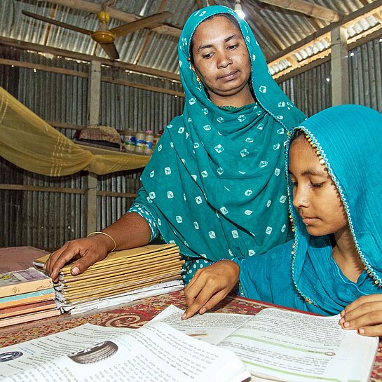Zwei Frauen mit grünen Kopftüchern beugen sich über einen Tisch mit Büchern und Schreibmaterial