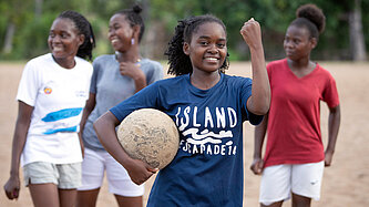 Eine junge Frau hält einen Fußball unterm Arm und die andere Hand zur Faust in die Luft gestreckt, hinter ihr stehen drei Mitspielerinnen