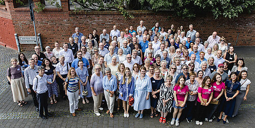 Großes Gruppenfoto der Teilnehmenden beim 8. Internationalen Stiftungstreffen der Stiftung Hilfe mit Plan 2026 in Königswinter, aufgenommen im Freien vor einer Backsteinmauer.