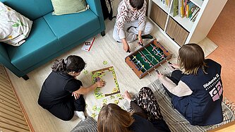 Zwei Mitarbeiterinnen einer Diakonie spielen in einer gemütlichen und hellen Unterkunft mit zwei Kindern. Der Raum ist wie ein Wohnzimmer eingerichtet und sie spielen auf einem Teppich auf dem Boden. 