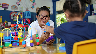 Eine Frau reicht einen blauen Bauklotz an ein junges Mädchen.