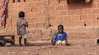 Ein Junge sitzt vor einem Lehmhaus, ein weiterer Junge steht neben ihm