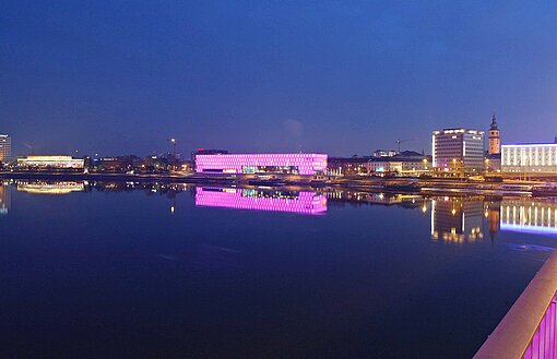 Inmitten einer abendlichen Stadtkulisse an einem Flussufer erstrahlt ein kastenartiges Gebäude in Pink