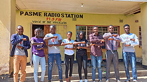 Gruppe junger Menschen posiert vor einer Radiostation in Sambia und stellen mit ihren Armen Gleichzeichen als Zeichen für Gleichberechtigung dar.