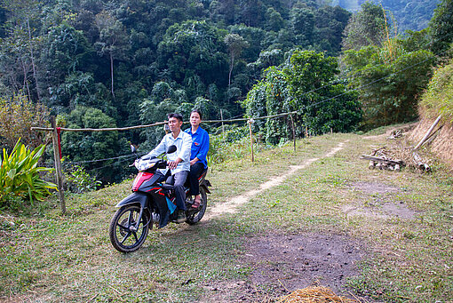 Zwei vietnamesische Personen sitzen auf einem Motorrad, um sie herum ist ländliche Natur