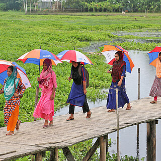 Frauen in bunten Kleidern und mit bunten Regenschirmen laufen über eine Brücke