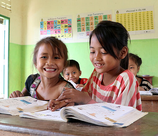 Lachende Schulkinder beim gemeinsamen Lernen in einer Grundschule in Laos. Zwei lachende Mädchen sitzen mit offenen Schulbüchern in einem Klassenzimmer in Laos und lernen gemeinsam.