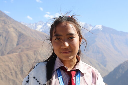 Portrait einer Jugendlichen, die in die Kamera lächelt. Im Hintergrund sieht man die Berge des Himalayas