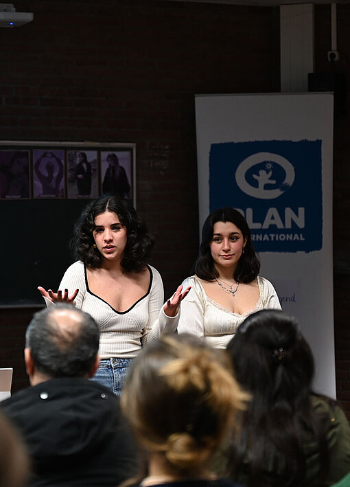 Zwei junge Frauen stehen in einem kleinen Hörsaal und sprechen zum Publikum.