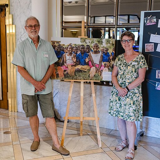 Ein Mann und eine Frau stehen im Foyer eines Veranstaltungsortes neben einer Foto-Ausstellung mit Kindern. Im Hintergrund ist eine Pinnwand mit Nachrichten und Bildern zu sehen, aufgenommen beim Stiftungstreffen 2026.