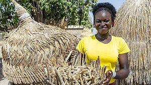 Junge Frau in gelbem Shirt präsentiert einen Korb mit geernteter Hirse vor traditionellen Strohhütten in Westafrika.