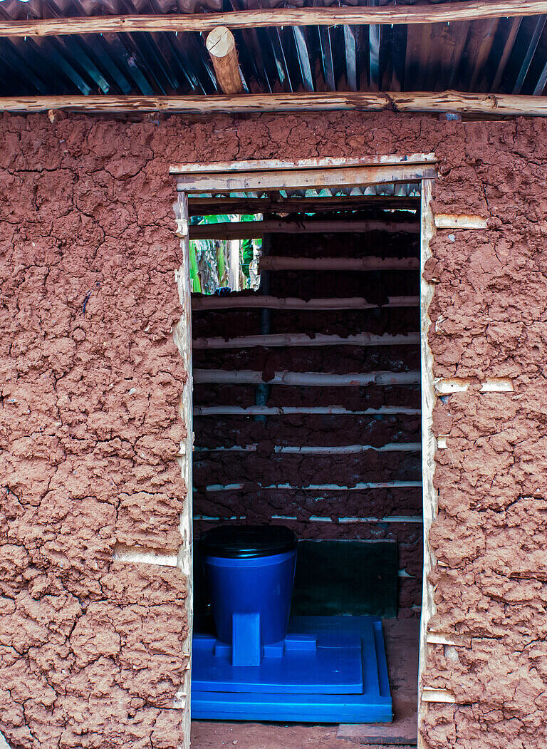 Ein Toilettenhäuschen aus Lehm, im inneren sieht man eine Toilette aus blauem Kunststoff