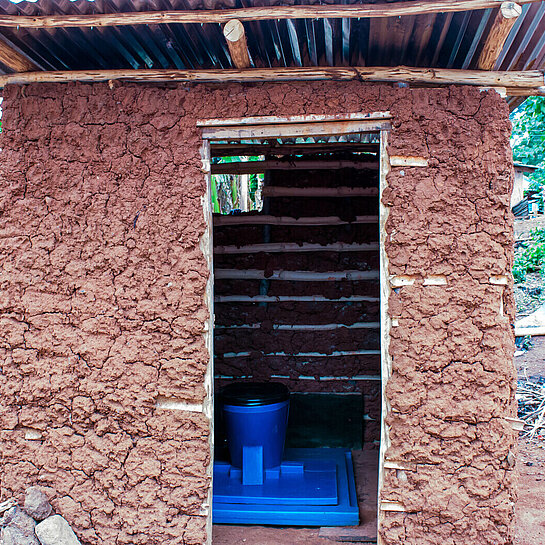 Ein Toilettenhäuschen aus Lehm, im inneren sieht man eine Toilette aus blauem Kunststoff