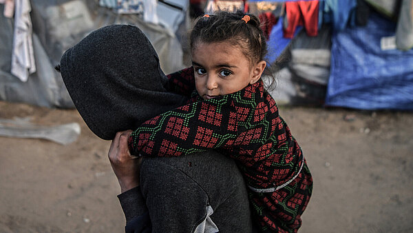 Ein Mädchen, das im Flüchtlingslager in Rafah im südlichen Gazastreifen lebt, wird von einer Person auf dem Rücken getragen. Sie schaut direkt in die Kamera. Im Hintergrund sind Zelte und Planen.