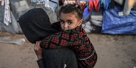 Ein Mädchen, das im Flüchtlingslager in Rafah im südlichen Gazastreifen lebt, wird von einer Person auf dem Rücken getragen. Sie schaut direkt in die Kamera. Im Hintergrund sind Zelte und Planen.