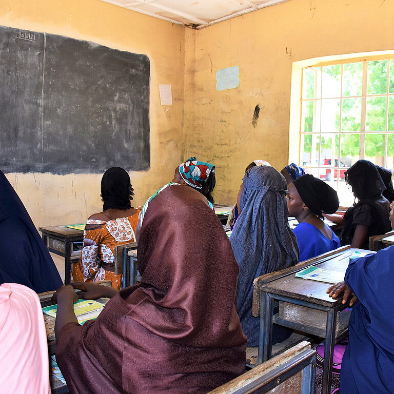 Ansicht einer Schulklasse in Nigeria, die Schülerinnen sind von hinten zu sehen und blicken auf die Tafel