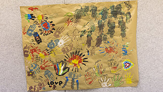 Ein großes Plakat voller bunter Handabdrücke und Zeichnungen von Kindern hängt an einer Wand.