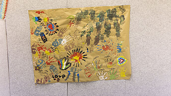 Ein großes Plakat voller bunter Handabdrücke und Zeichnungen von Kindern hängt an einer Wand.