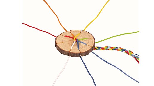 Strickscheibe im Einsatz. Verschiedenenfarbige Fäden sind eingefäldet und die bunte fertige Kordel lschut unterhalb hervor.