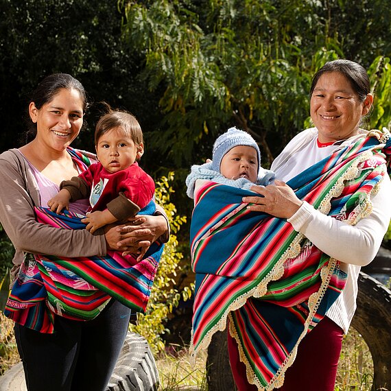 Zwei Frauen stehen nebeneinander im Freien und tragen jeweils ein Baby in bunten Tüchern vor der Brust; im Hintergrund stehen Bäume und große Reifen.