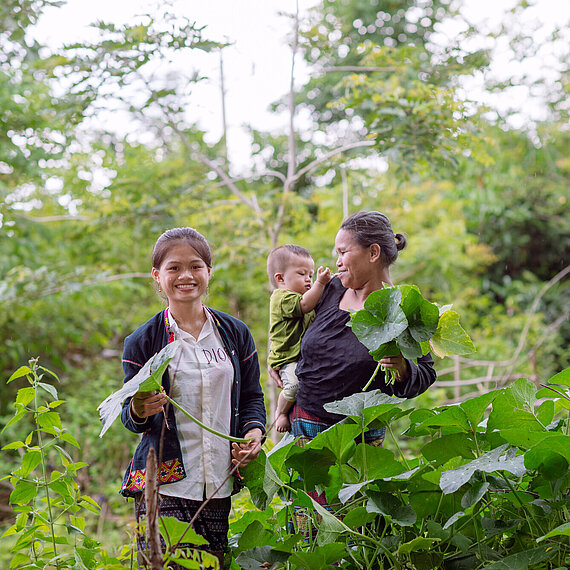 Zwei Frauen und ein Kleinkind stehen inmitten grüner Pflanzen. Die Frauen lächeln und halten große Blätter in der Hand. Im Hintergrund ist dichte Vegetation und Wald zu sehen.