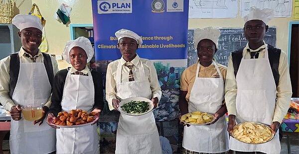 Fünf Jugendliche in Schuluniformen tragen weiße Kochschürzen und -mützen und stehen in einem Klassenraum. Jede Person hält einen Teller mit zubereiteten Speisen, im Hintergrund hängt ein Banner von Plan International.