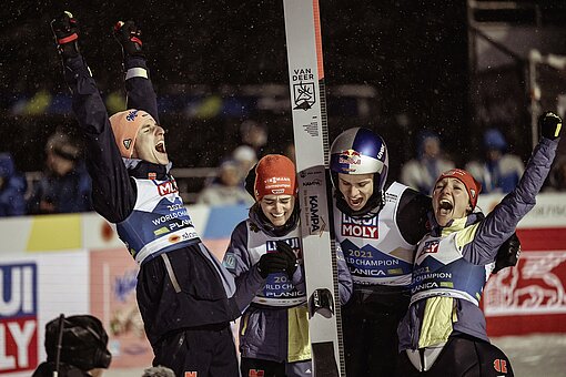 Zwei Skispringer und zwei Skispringerinnen stehen nebeneinander, halten ihre Skier und jubeln