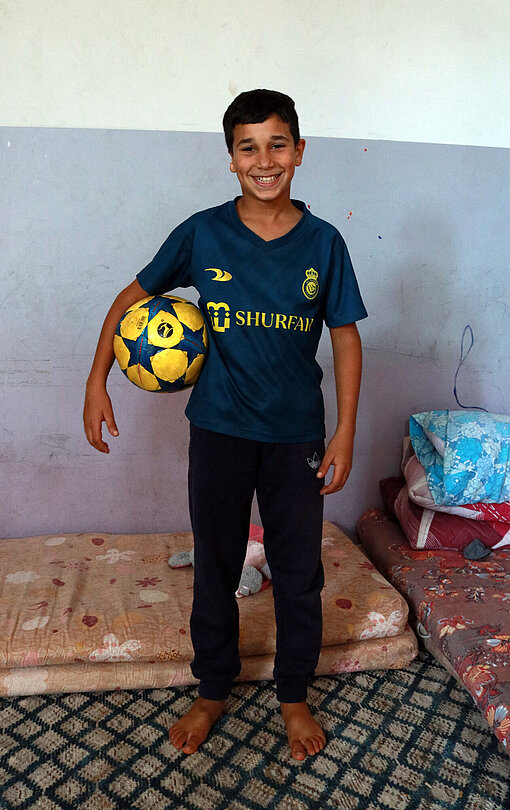 Ein Junge steht barfuß vor einer Matratze, die auf dem Boden liegt, er hat einen Fußball unter den rechten Arm geklemmt und lächelt