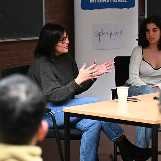 Maryam Blumenthal sitzt an einem Tisch in einem kleinen Hörsaal vor einem Plan-Aufsteller.
