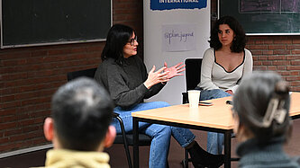 Maryam Blumenthal sitzt an einem Tisch in einem kleinen Hörsaal vor einem Plan-Aufsteller.
