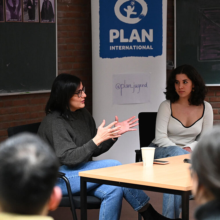 Maryam Blumenthal sitzt an einem Tisch in einem kleinen Hörsaal vor einem Plan-Aufsteller.