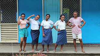 Fünf Mädchen oder junge Frauen aus den Salomonen stehen selbstbewusst und lachend vor einem blauen Schulgebäude; einige tragen Schuluniformen, andere Freizeitkleidung.