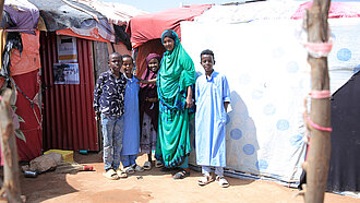 Eine Frau, ein Mädchen und drei jungen stehen vor einer Behelfsunterkunft aus Stoffen und Platten. 