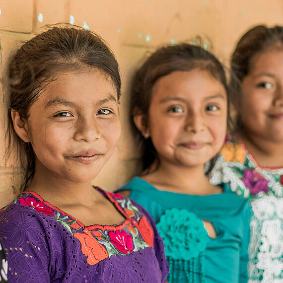 Drei Mädchen in traditioneller Kleidung lächeln in die Kamera – aufgenommen in einer Schule in Guatemala