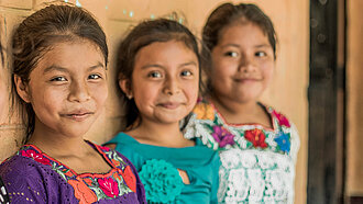 Drei Mädchen in traditioneller Kleidung lächeln in die Kamera – aufgenommen in einer Schule in Guatemala